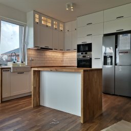 Lagos Meble - Jasna kuchnia z białymi szafkami i drewnianym blatem, wyspa kuchenna z drewnianymi bokami, widoczny zlew farmerski i nowoczesna lodówka.