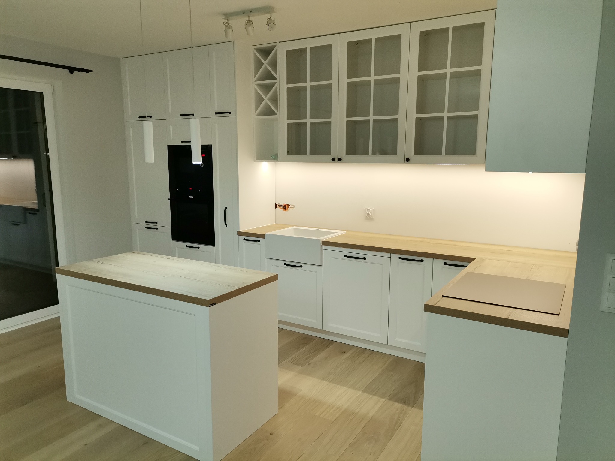 Jasna kuchnia z białymi szafkami, drewnianymi blatami i wyspą kuchenną, oświetlona listwą LED pod szafkami wiszącymi, z czarnymi uchwytami i zabudowanym piekarnikiem.