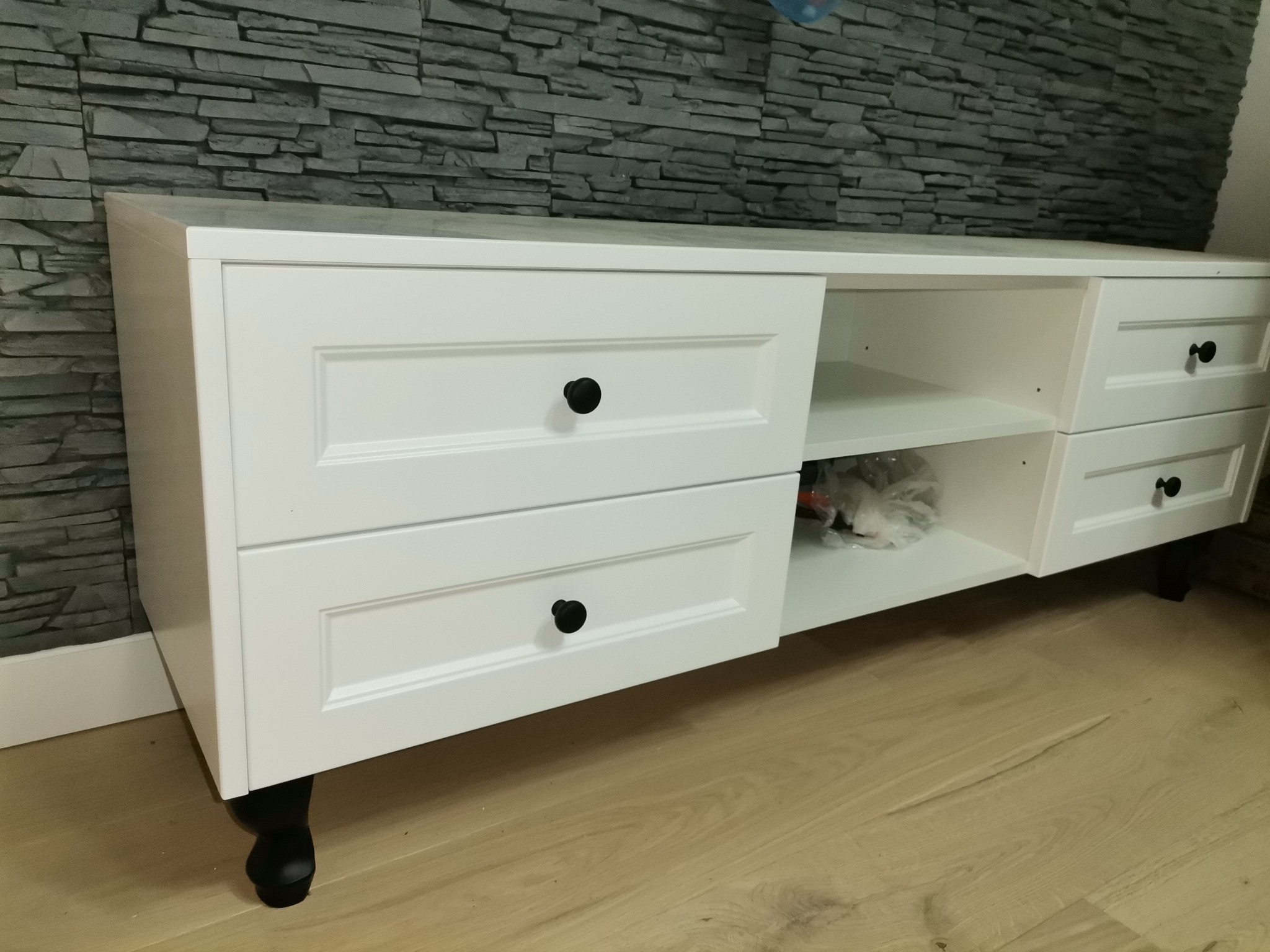 Biała szafka RTV z szufladami i otwartą półką, czarne okrągłe uchwyty, dekoracyjna ściana z kamienia w tle, drewniana podłoga.
