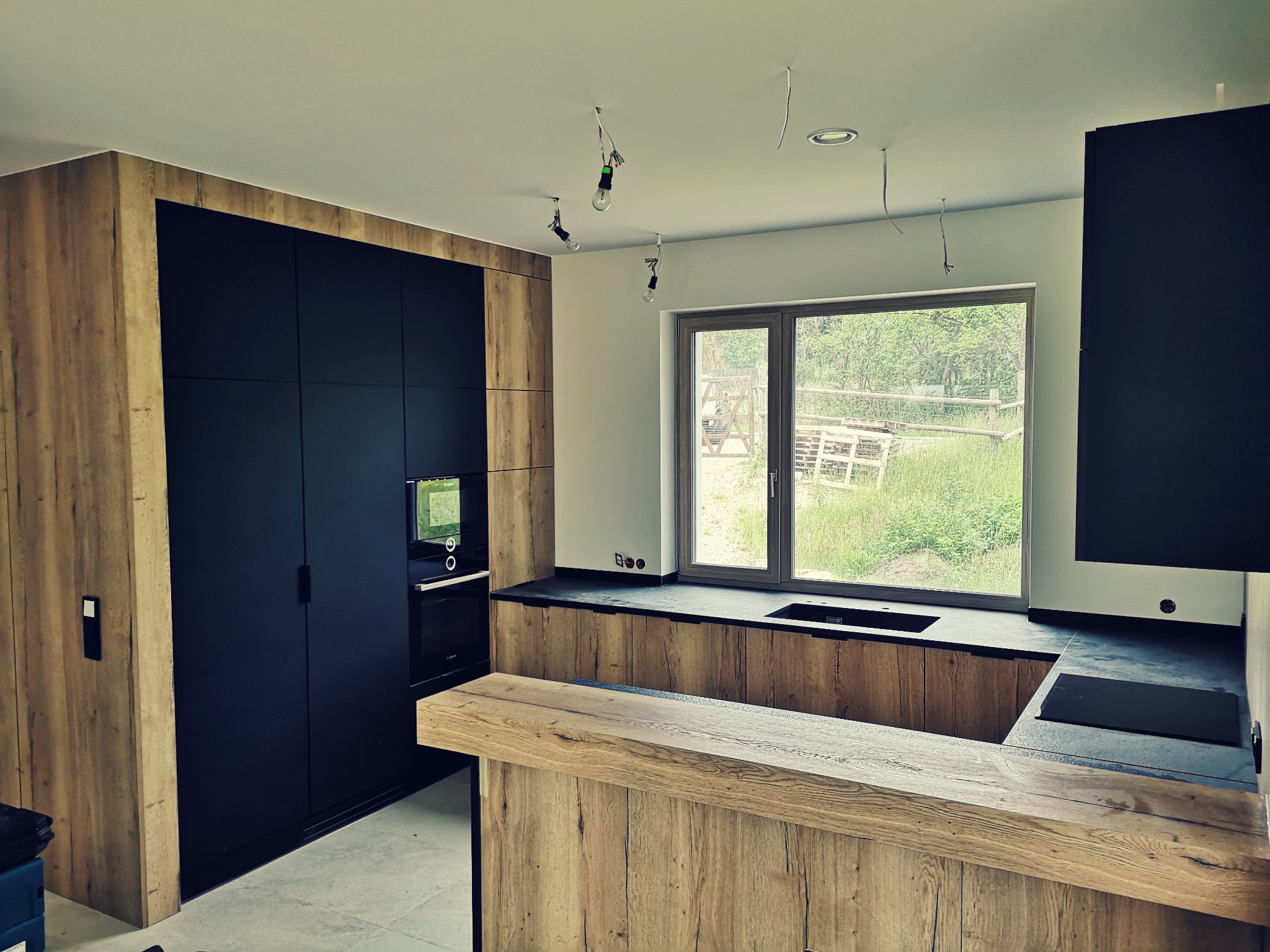 Nowoczesna kuchnia z czarnymi frontami szafek i drewnianymi elementami wykończeniowymi, widoczne niedokończone oświetlenie sufitowe i okno z widokiem na zewnątrz.