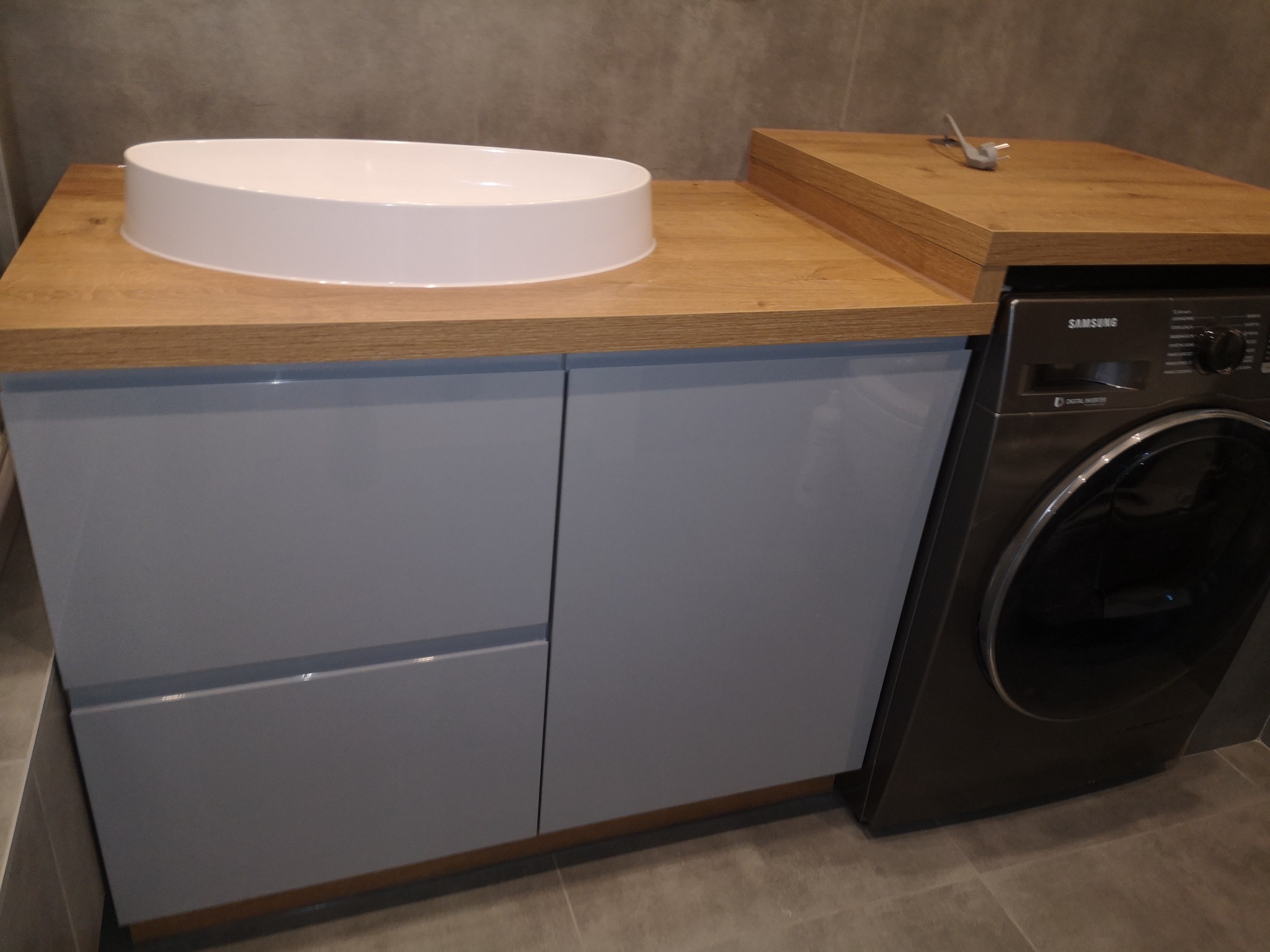 Łazienka z umywalką nablatową na szafce z szarymi frontami, obok pralka Samsung z blatem drewnianym, szary kabel zasilający na blacie