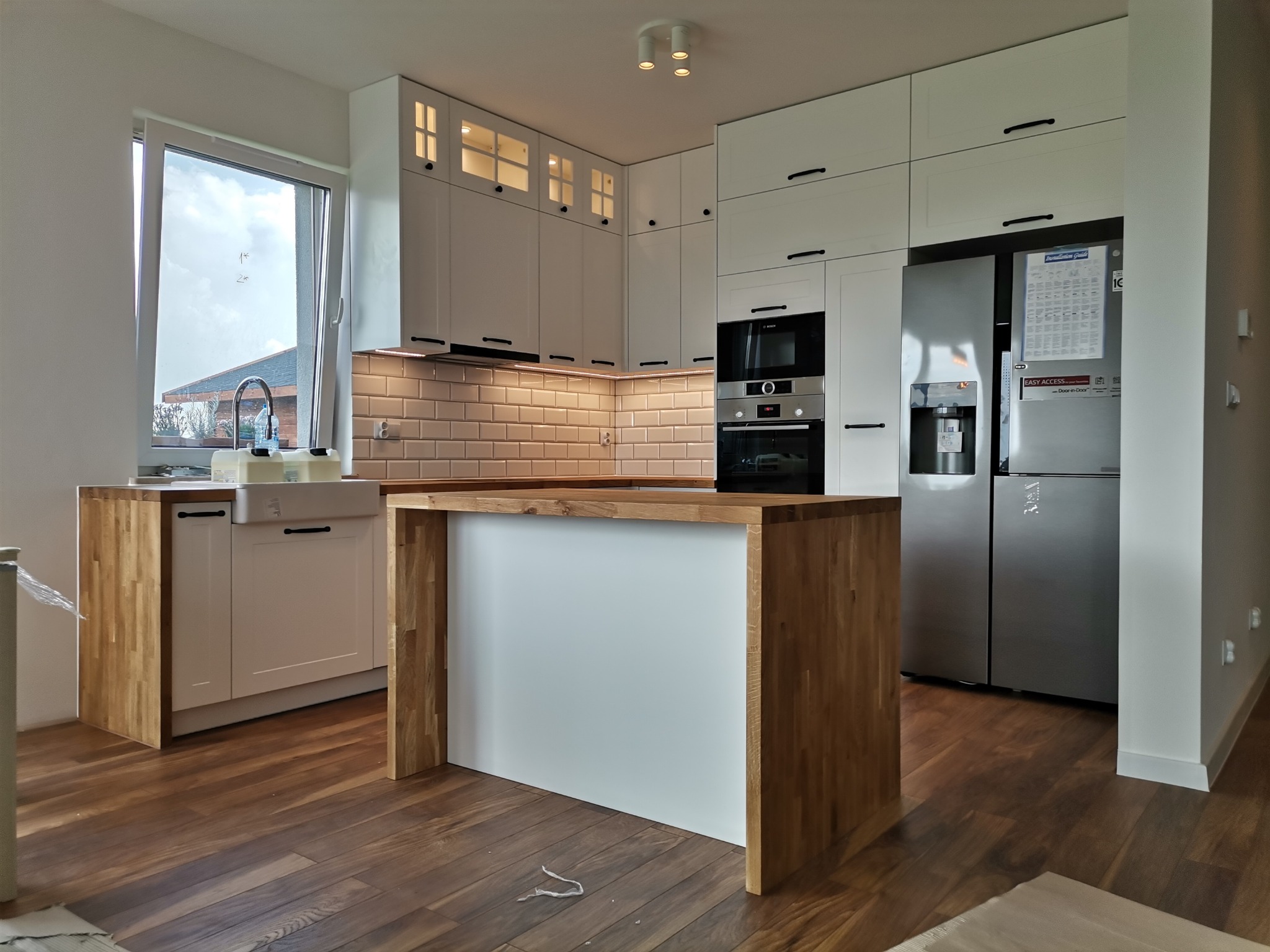 Jasna kuchnia z białymi szafkami i drewnianym blatem, wyspa kuchenna z drewnianymi bokami, widoczny zlew farmerski i nowoczesna lodówka.