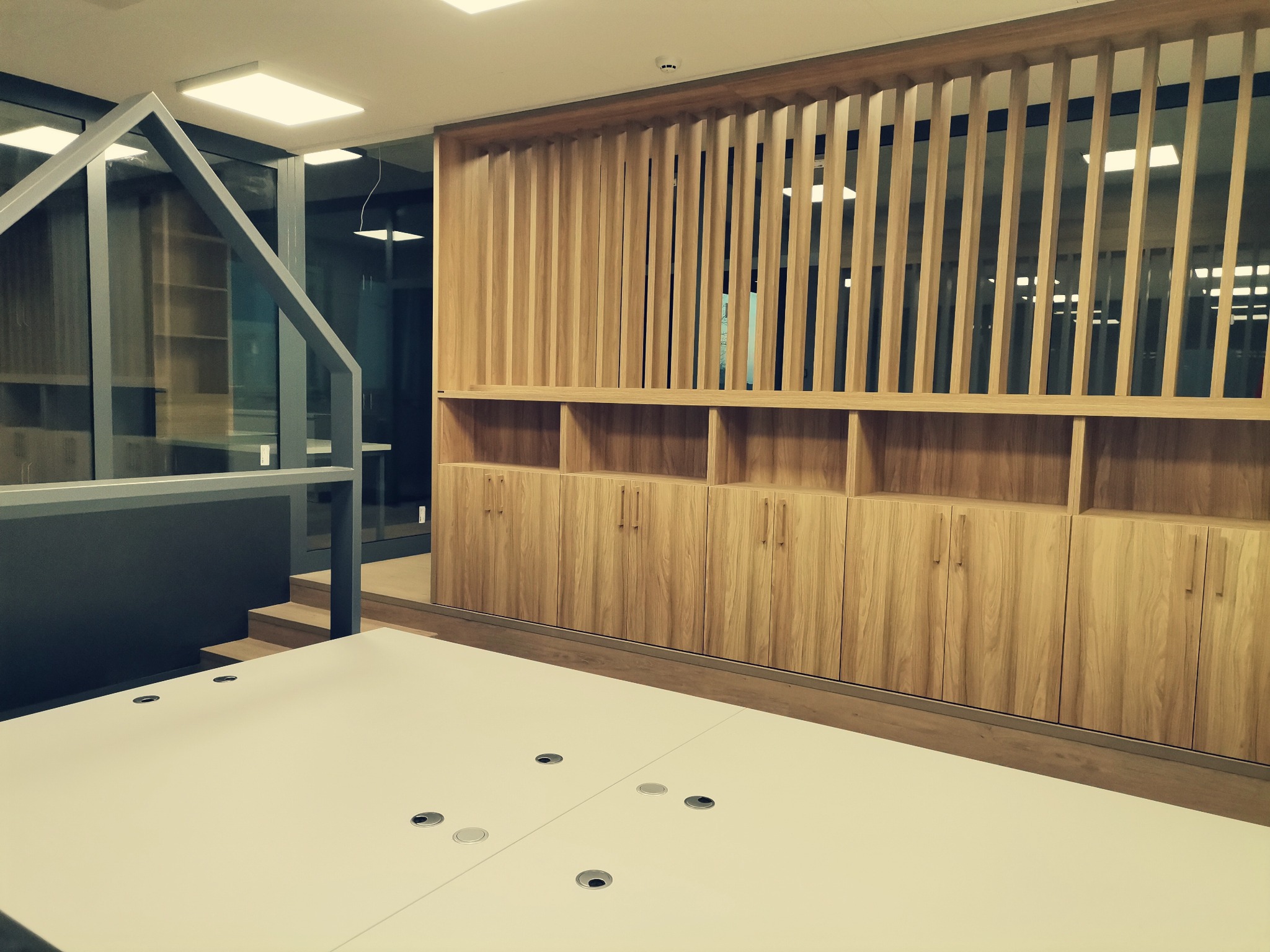 Nowoczesne biuro z drewnianą zabudową meblową: szafki z półkami i ażurowa ścianka działowa z pionowych listew, białe biurka z otworami na kable.