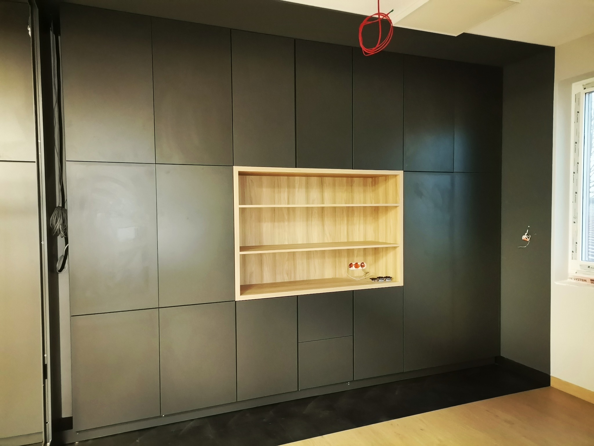 Ciemnoszara zabudowa meblowa z otwartą, jasną półką z drewna we Wrocławiu, widoczne niedokończone podłączenia elektryczne i gniazdka.