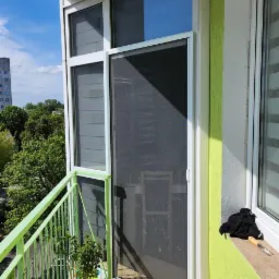 Balkon z zieloną balustradą, na którym zamontowano przesuwną moskitierę w białej ramie, obok okna z roletami wewnętrznymi, z widocznymi doniczkami z kwiatami.