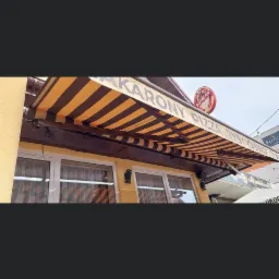 Zewnętrzna, brązowo-żółta, pasiakowa osłona przeciwsłoneczna nad oknami lokalu gastronomicznego z widocznym szyldem reklamowym.