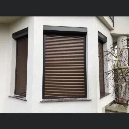 Brązowe osłony okienne zewnętrzne zamontowane w białej fasadzie budynku, widoczne trzy okna z zamkniętymi listwami.