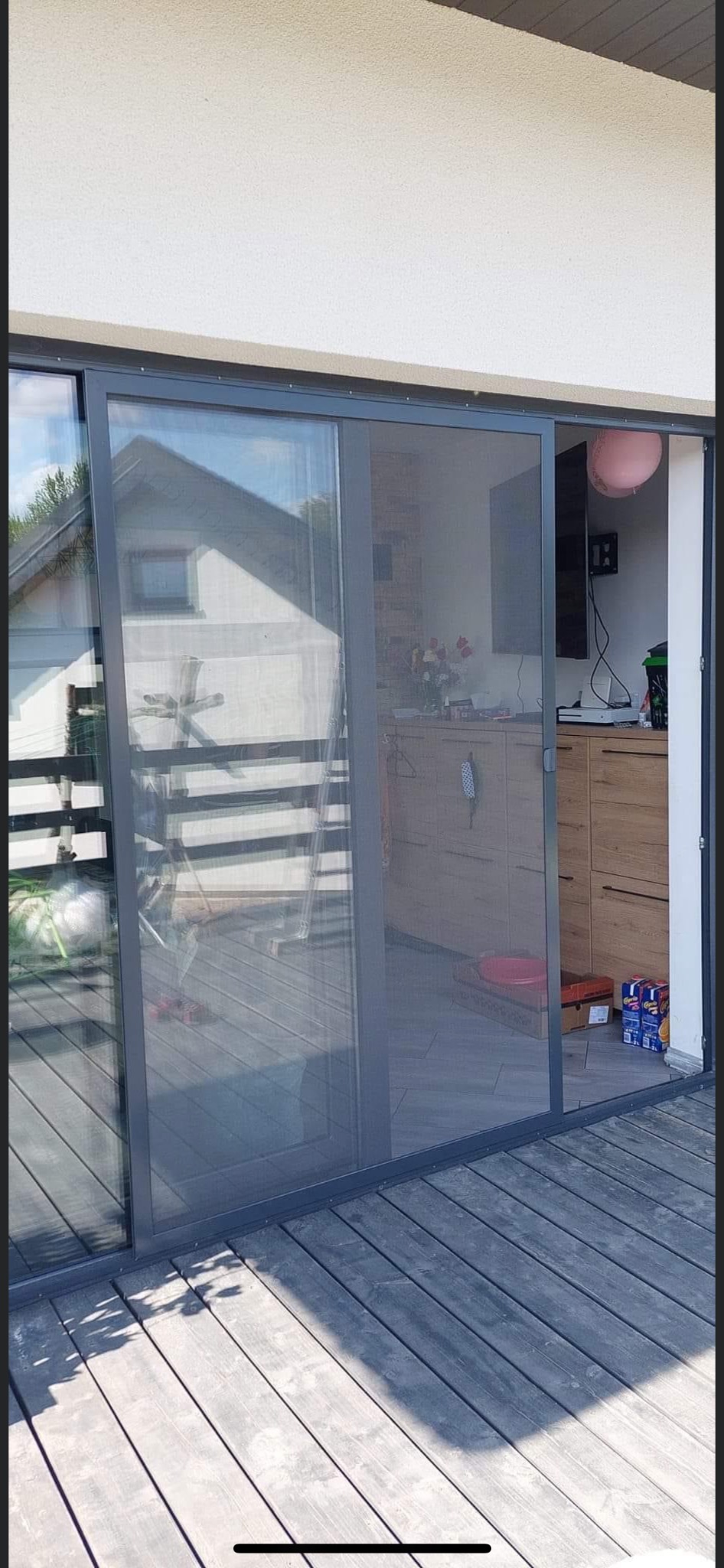 Przesuwne drzwi tarasowe z szarą ramą i zamontowaną moskitierą, widok z zewnątrz na drewniany taras.