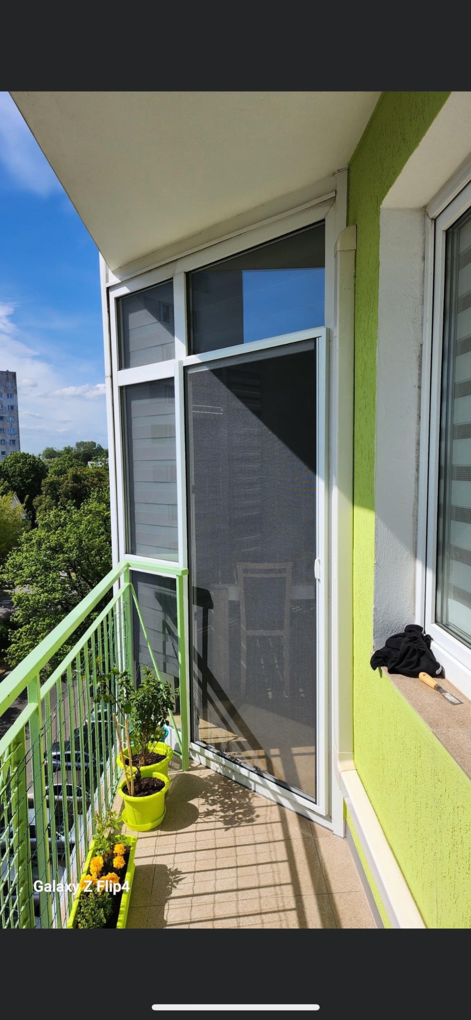 Balkon z zieloną balustradą, na którym zamontowano przesuwną moskitierę w białej ramie, obok okna z roletami wewnętrznymi, z widocznymi doniczkami z kwiatami.