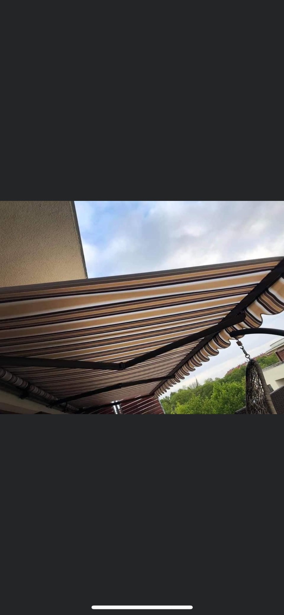 Rozwinięta markiza w odcieniach beżu i brązu, z falbaną, zamontowana na balkonie, widok z dołu na niebo i zieleń drzew w tle.