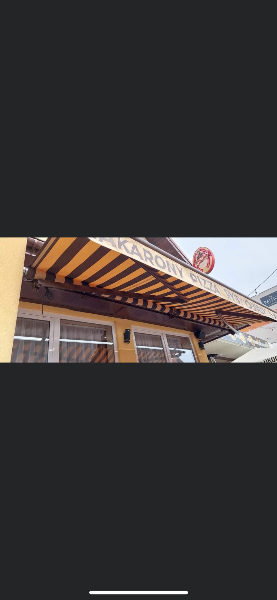 Zewnętrzna, brązowo-żółta, pasiakowa osłona przeciwsłoneczna nad oknami lokalu gastronomicznego z widocznym szyldem reklamowym.