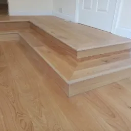 Wykończone drewniane schody wewnętrzne, z widocznymi detalami połączeń i słojów drewna, prowadzące do białych drzwi.