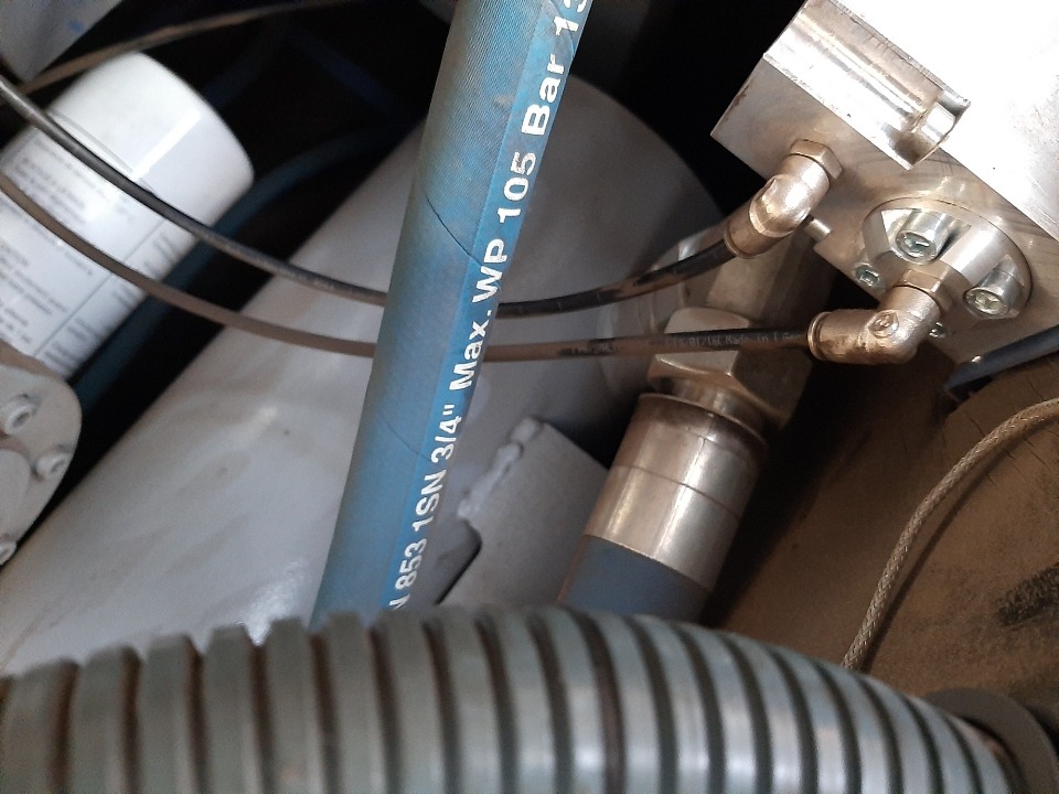 Szczegółowe ujęcie z bliska na podzespoły maszyny: niebieski wąż z napisem 'Max. WP 105 Bar 13', metalowe złączki i przewody, filtr cylindryczny oraz karbowana rura ochronna.