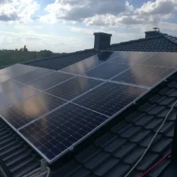 Panele fotowoltaiczne zamontowane na czarnym, spadzistym dachu z dachówki ceramicznej w słoneczny dzień, widoczne kominy i liny zabezpieczające.