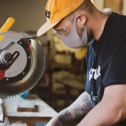 Stolarz w czarnej koszulce z tatuażem na ramieniu, żółtej czapce i ochronnej masce na twarzy, tnący drewno za pomocą piły tarczowej. Nosi okulary ochronne i zegarek na bransolecie.