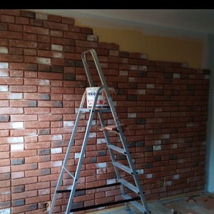 Ściana wyłożona dekoracyjną cegłą w odcieniach czerwieni i brązu, częściowo ukończona, z widoczną puszką farby i aluminiową drabiną opartą o ścianę. Fragment ściany powyżej cegieł pomalowany na żółto.