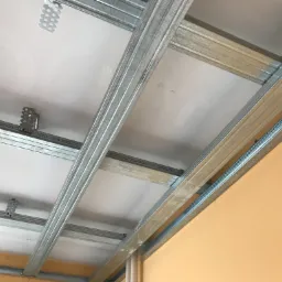 Montaż sufitu podwieszanego z płyt kartonowo-gipsowych, widoczna metalowa konstrukcja nośna i częściowo zamontowane płyty, jasna ściana w tle.