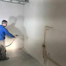 Mężczyzna w niebieskiej bluzie i roboczych spodniach aplikuje gładź natryskową na ścianę w pomieszczeniu w stanie deweloperskim, widoczne puszki elektryczne i metalowy stelaż sufitowy.