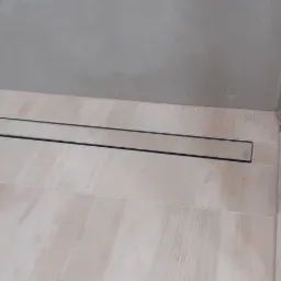 Nowoczesny odpływ liniowy w kabinie prysznicowej, jasne płytki podłogowe imitujące drewno, szklana ściana kabiny z chromowanymi okuciami.