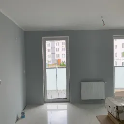 Wnętrze pomalowanego na szaro pokoju z białymi listwami przypodłogowymi, marmurową podłogą i grzejnikiem; widoczne okno balkonowe z widokiem na budynek mieszkalny i zapakowane kartony na podłodze.