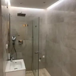 Nowoczesna łazienka z kabiną prysznicową ze szklanymi drzwiami, umywalką na szafce i podświetleniem LED na suficie, wykończona płytkami w odcieniach beżu i szarości.