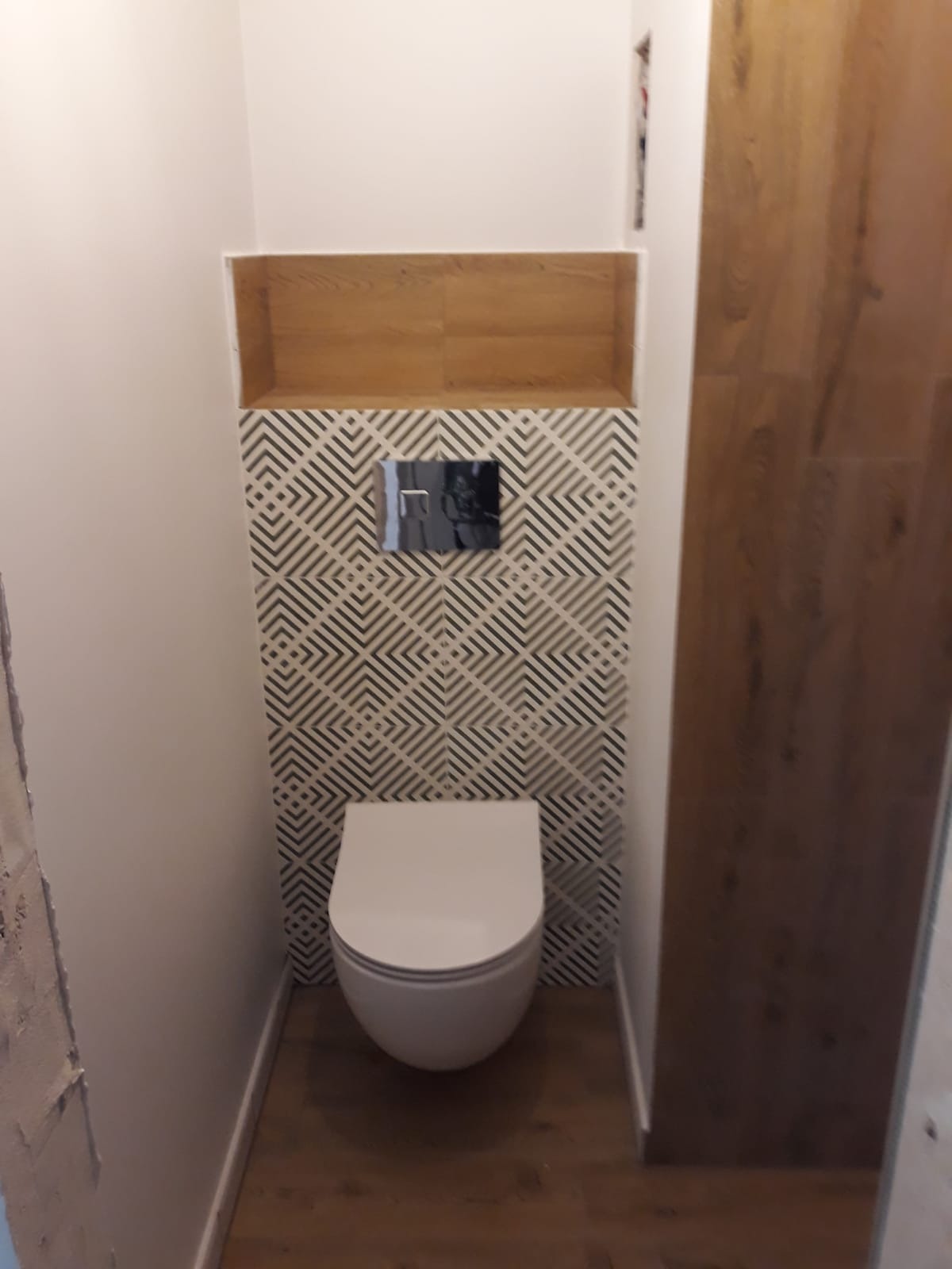 Wisząca toaleta w małej łazience, nad nią drewniana półka i wzorzyste płytki, na podłodze panele imitujące drewno.