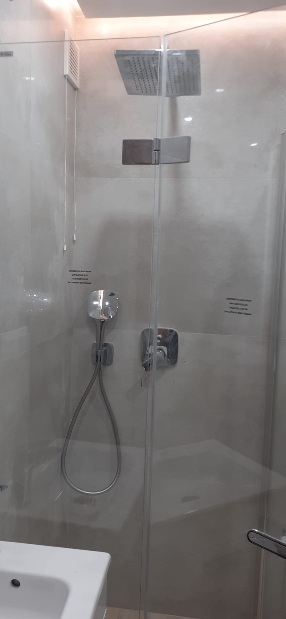 Nowoczesna kabina prysznicowa z deszczownicą i szklanymi drzwiami, widoczna bateria termostatyczna i biała umywalka w rogu.