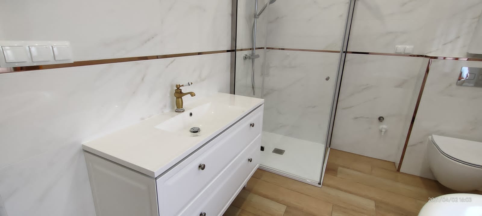 Nowoczesna łazienka z białymi marmurkowymi płytkami, kabiną prysznicową typu walk-in, białą szafką pod umywalkę z złotą baterią i drewnopodobną podłogą.