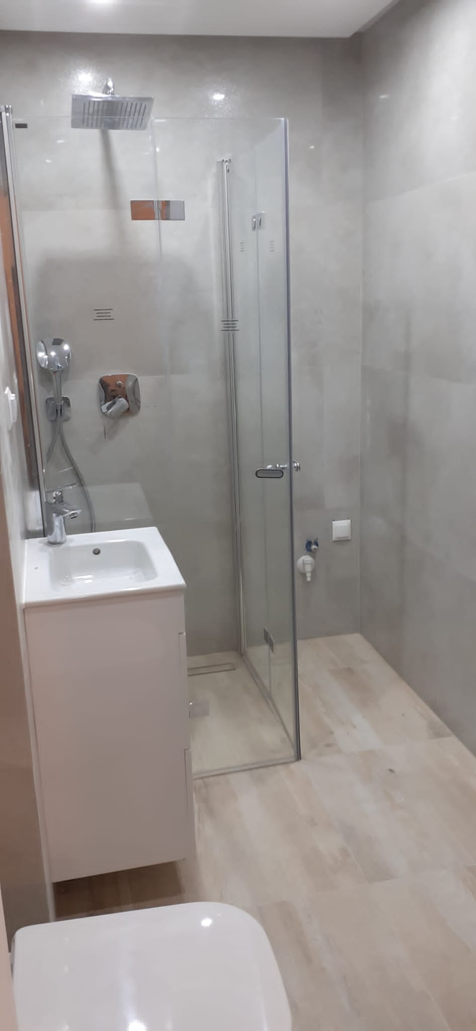 Wykończona łazienka z kabiną prysznicową typu walk-in z odpływem liniowym, umywalką z szafką w kolorze białym i armaturą chromowaną na tle beżowych płytek.