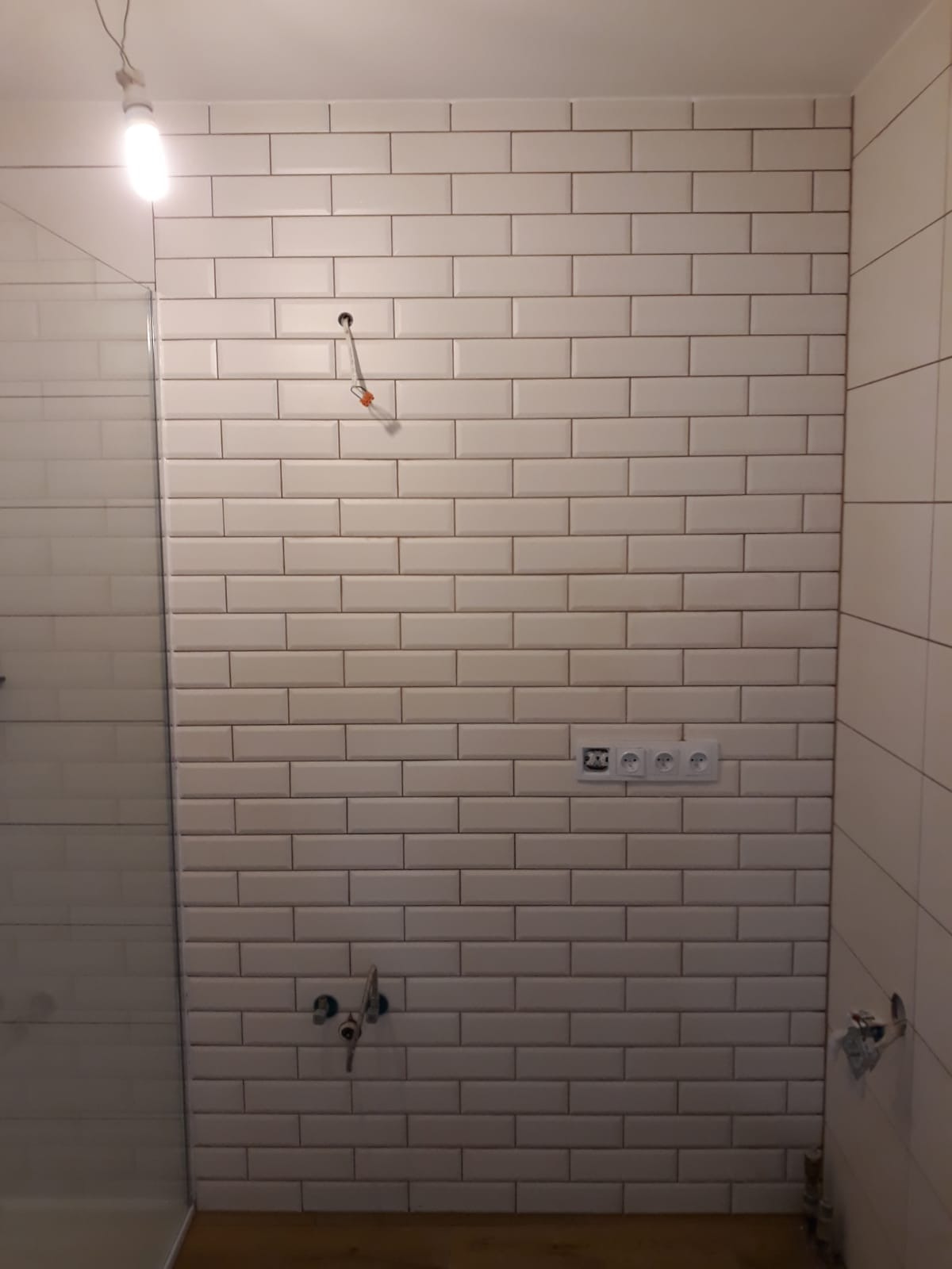 Ściana w łazience wyłożona białymi, podłużnymi płytkami ceramicznymi, widoczne gniazdka elektryczne, wyprowadzone przewody oświetleniowe i instalacja wodna przed montażem armatury.