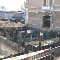 NOWER Budownictwo Barbara Nowak w spadku - Budowa fundamentów budynku obok torów kolejowych; widoczne szalunki, zbrojenia, robotnicy i przejeżdżający pociąg.