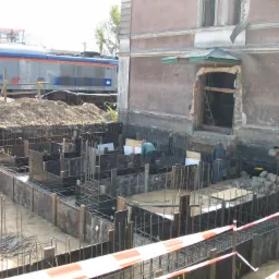 Budowa fundamentów budynku obok torów kolejowych; widoczne szalunki, zbrojenia, robotnicy i przejeżdżający pociąg.