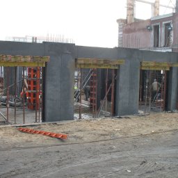 NOWER Budownictwo Barbara Nowak w spadku - Surowy stan budowy: betonowe ściany z otworami na okna, widoczne szalunki i zbrojenia, pracownicy w tle, pomarańczowa drabina leżąca na ziemi.