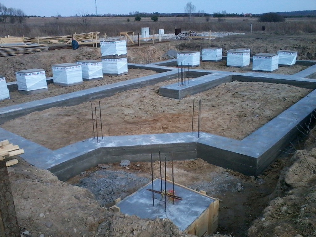 Wylane fundamenty domu jednorodzinnego z wystającymi prętami zbrojeniowymi, na działce widoczne materiały budowlane zabezpieczone folią.