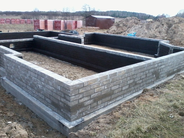 Wzniesione z cegły fundamenty budynku z hydroizolacją, na tle sterty cegieł i małego baraku.