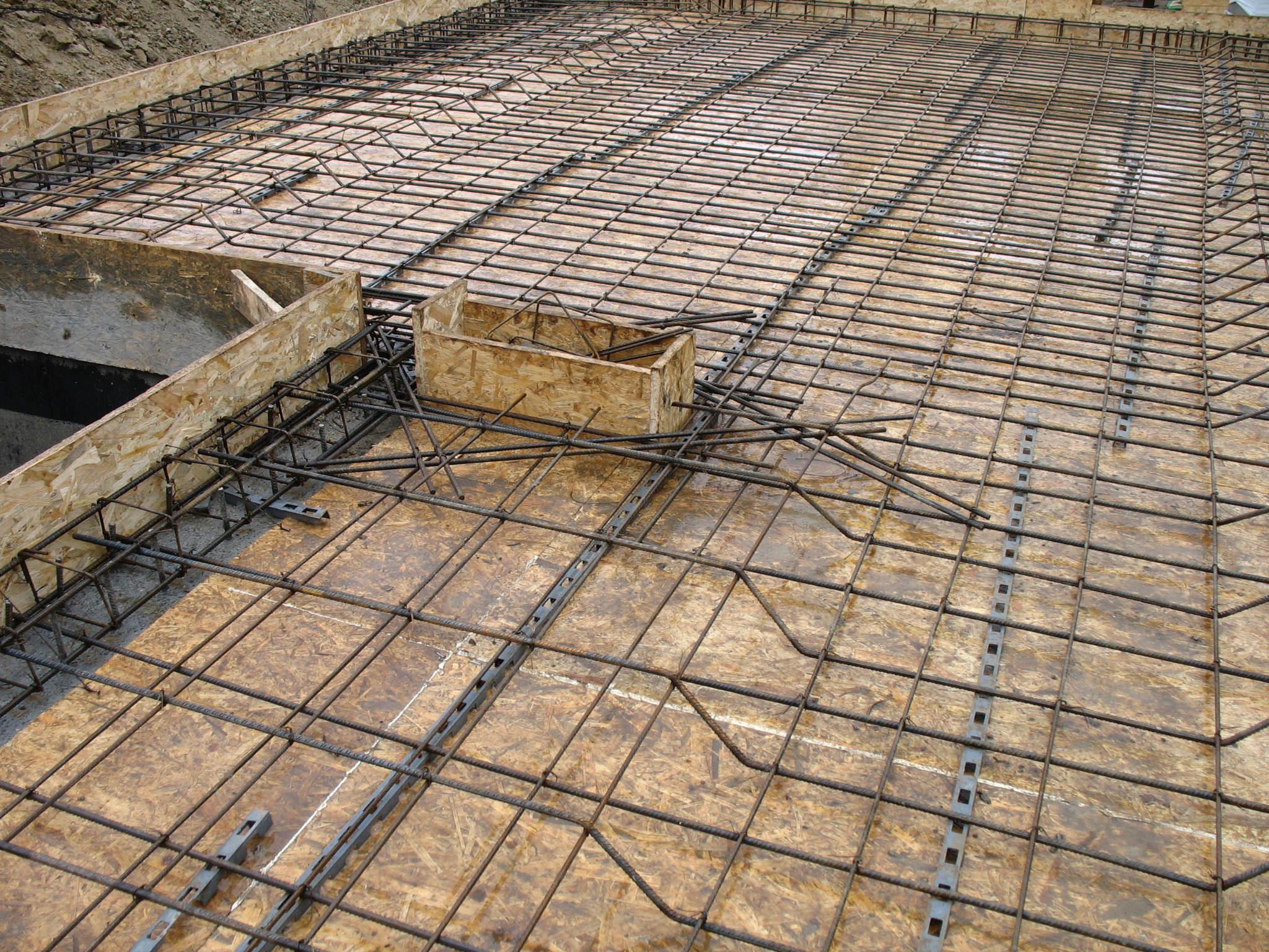 Zbrojenie płyty fundamentowej z deskowaniem i elementami dystansowymi, widoczne pręty zbrojeniowe ułożone w kratę na płycie ze sklejki.