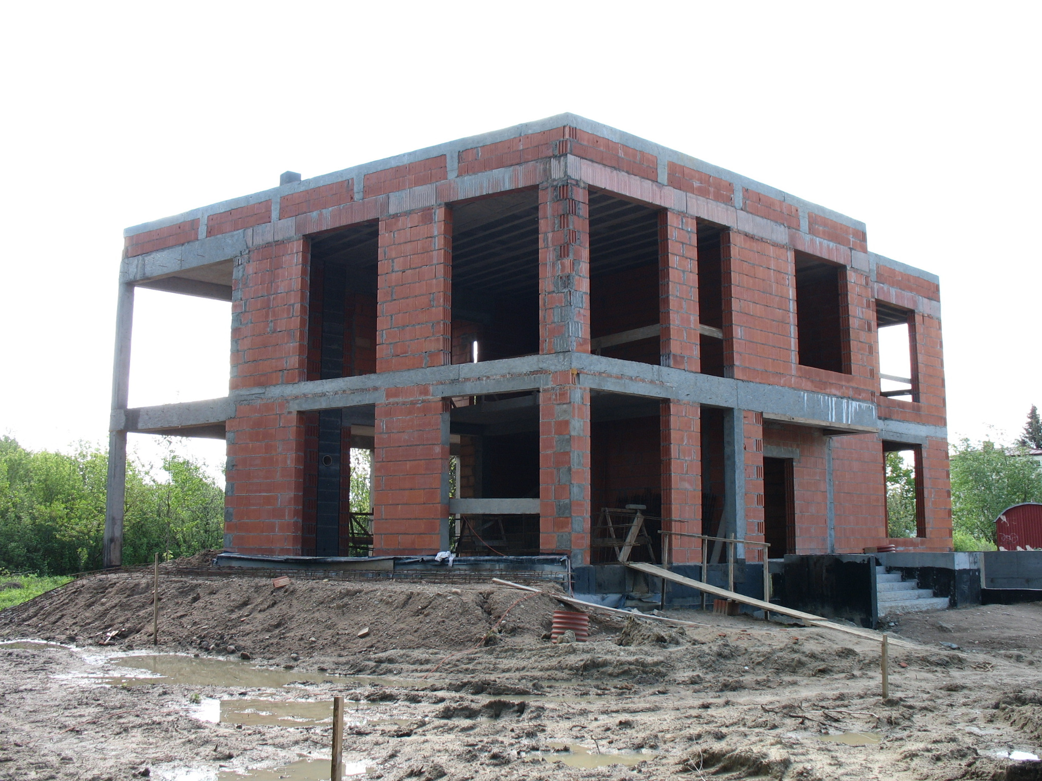 Dwukondygnacyjny budynek w stanie surowym otwartym, z czerwonej cegły i betonu, widoczny z niskiej perspektywy na tle zieleni i błotnistego terenu budowy.
