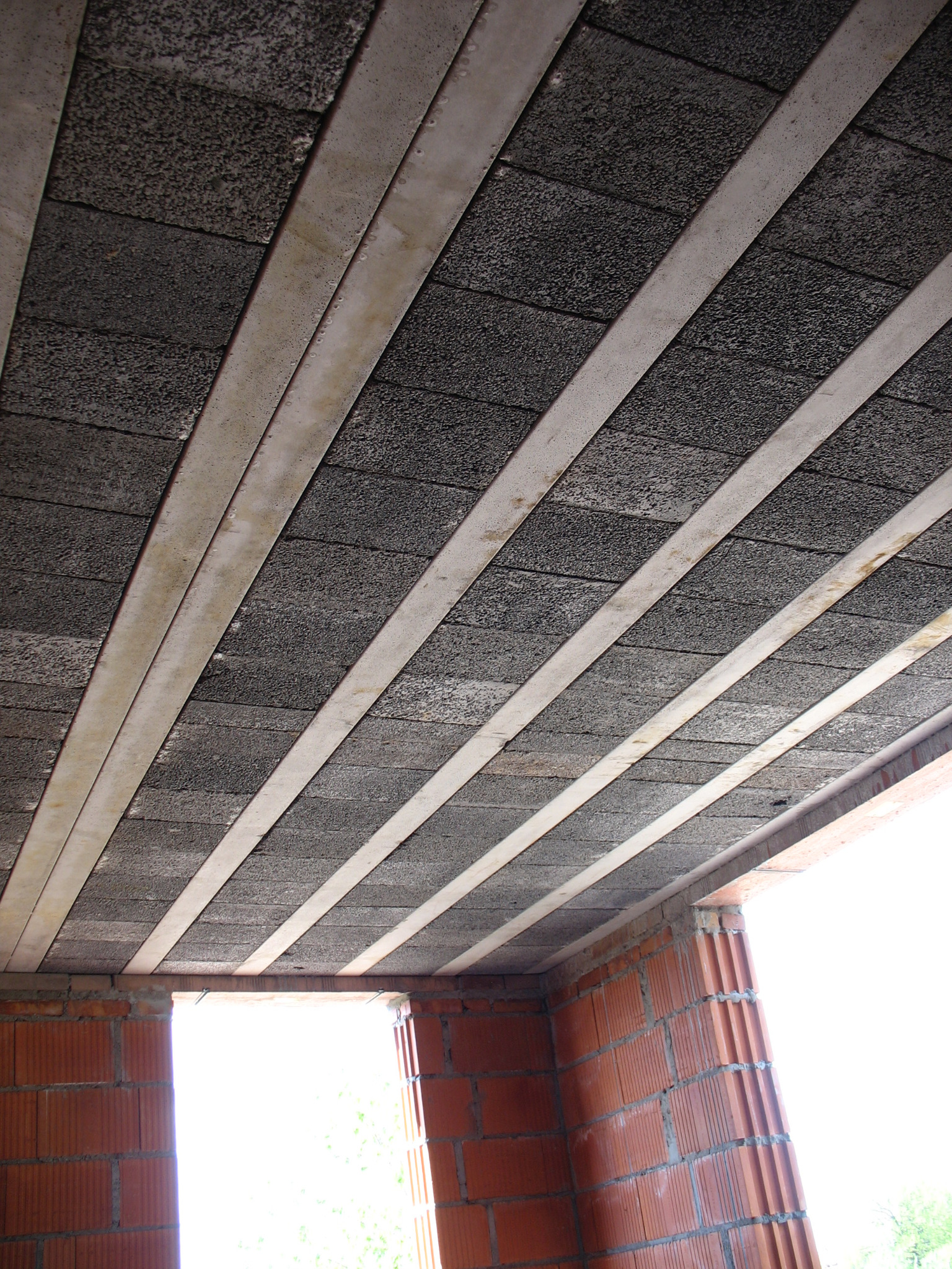 Wnętrze budynku w trakcie budowy: strop z szarych pustaków betonowych i białych belek nośnych, widok z dołu na surową konstrukcję i fragment muru z czerwonej cegły.