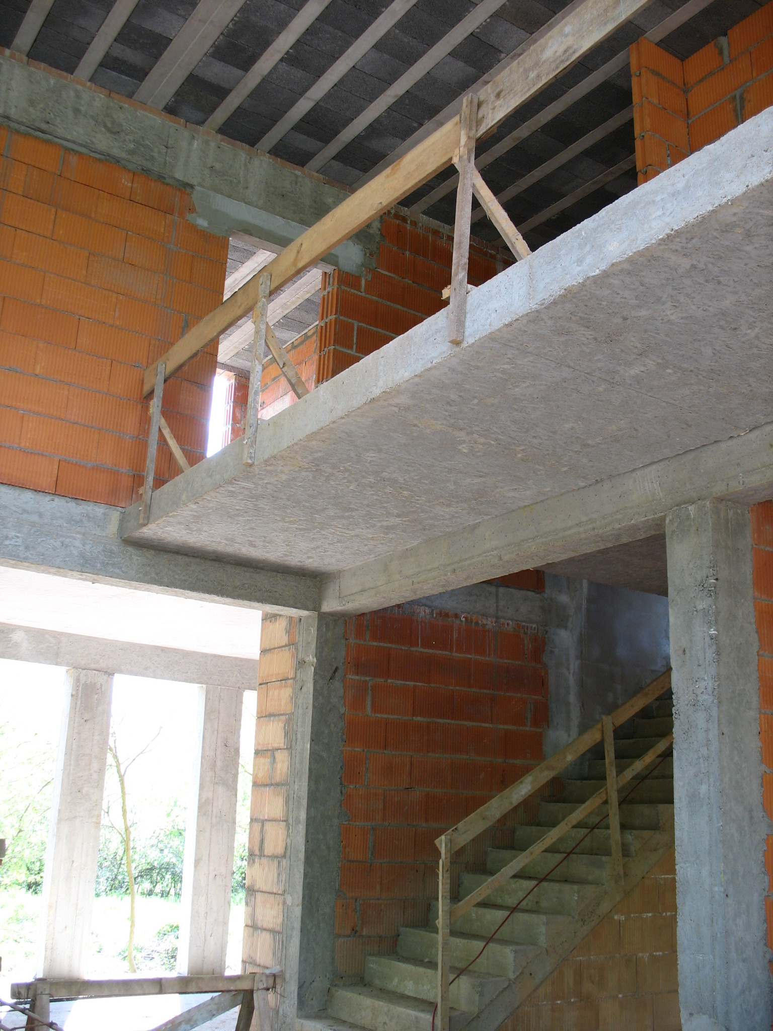 Surowy stan budynku w trakcie budowy: widoczne betonowe stropy, ceglane ściany z pustaków ceramicznych, drewniane barierki schodów i prowizoryczne zabezpieczenia na antresoli, sufit z betonowych płyt.