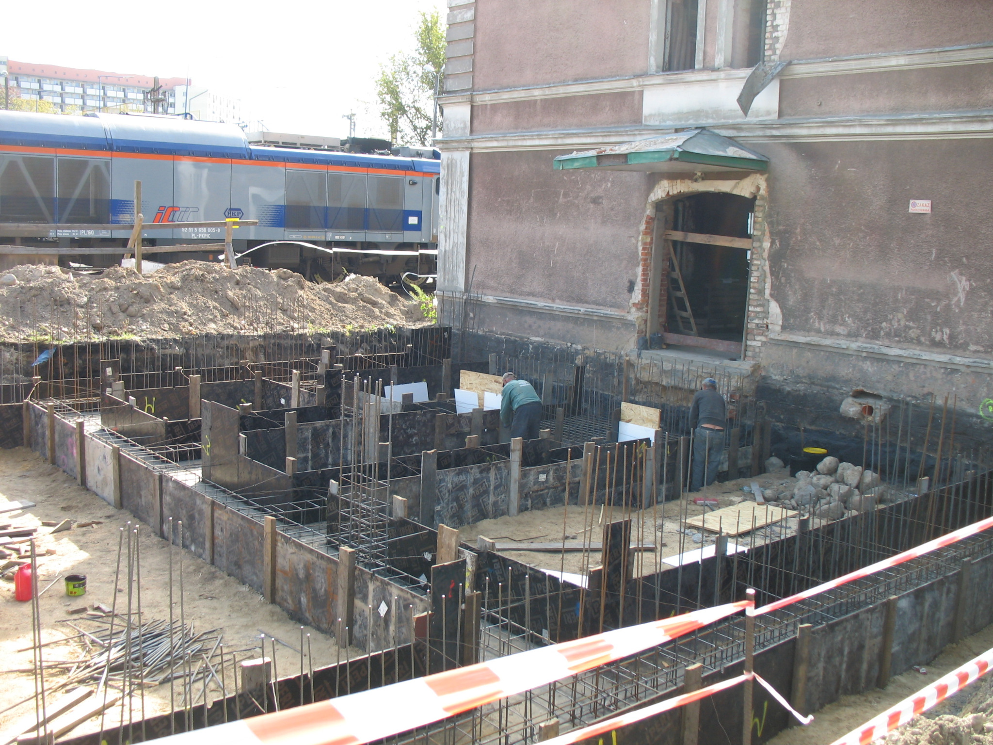 Budowa fundamentów budynku obok torów kolejowych; widoczne szalunki, zbrojenia, robotnicy i przejeżdżający pociąg.