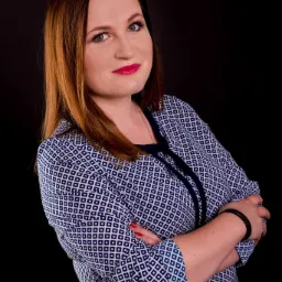 Portret kobiety z rudymi końcówkami włosów, ubranej w niebieską koszulę w białe romby, na czarnym tle. Skrzyżowane ramiona, czerwona szminka i ciemny skórzany pasek na nadgarstku.