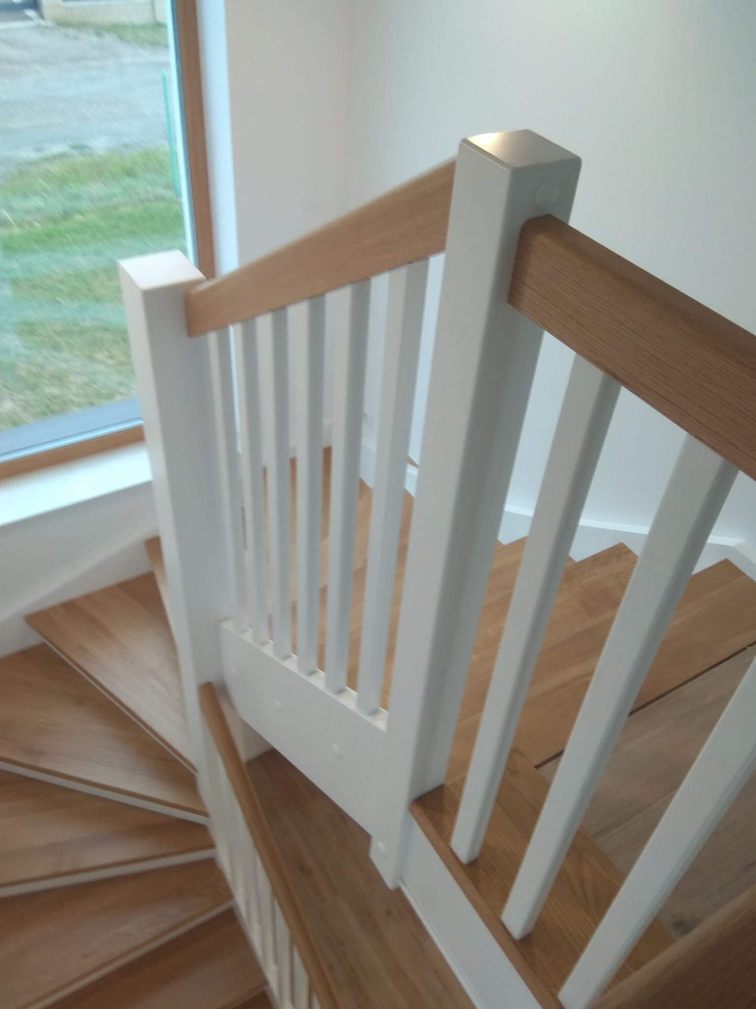 Biała balustrada z drewnianą poręczą i tralkami na spiralnych schodach w Zakliczynie, widok z góry na zakręt schodów.