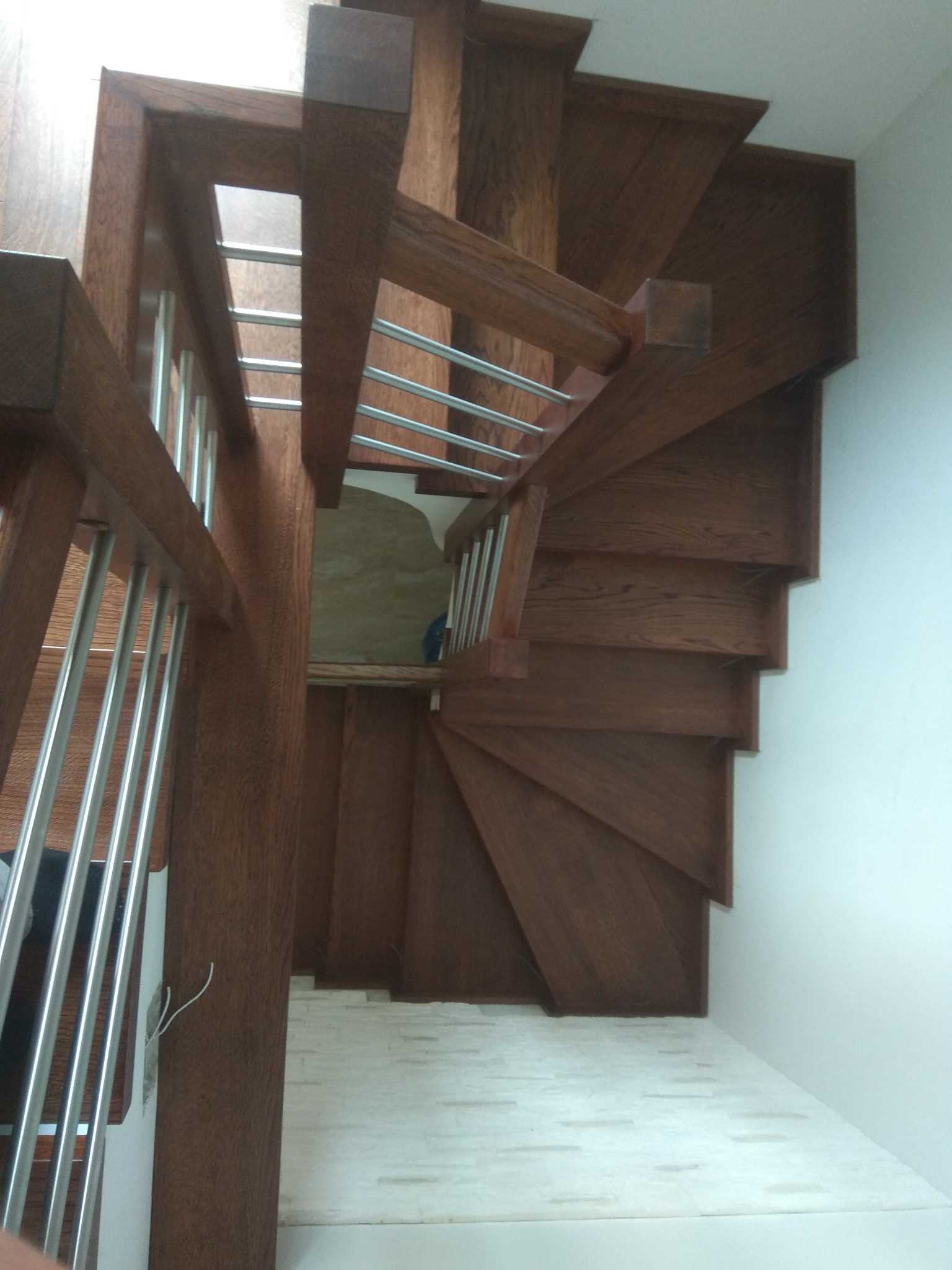 Wysoki kąt widzenia na spiralne schody z ciemnego drewna z metalowymi balustradami, widoczne białe ściany i podłoga z jasnych płytek.