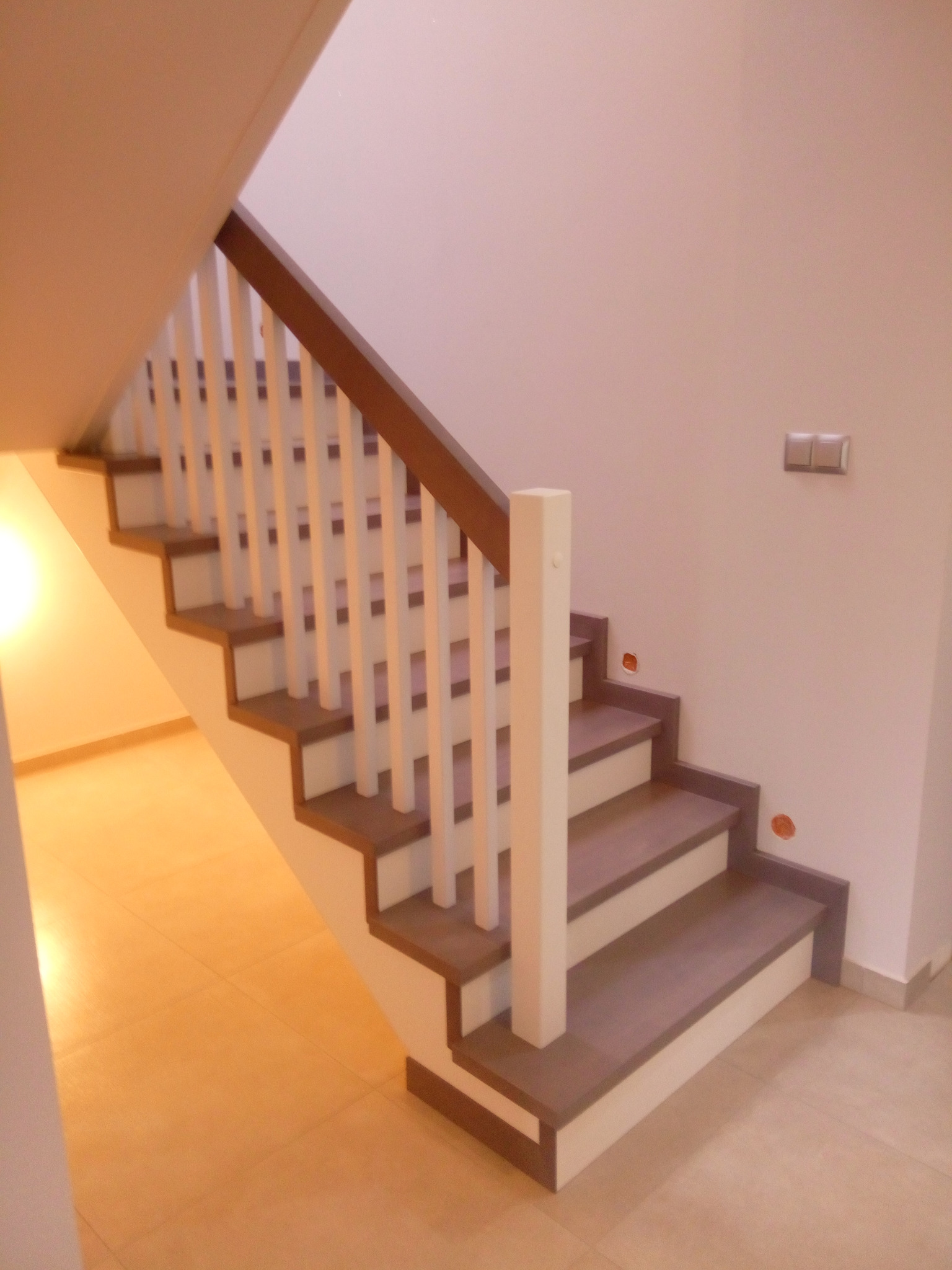 Widok na jasne schody z drewnianymi stopniami i białą balustradą z brązową poręczą, widoczne niedokończone otwory na gniazdka elektryczne na ścianie.