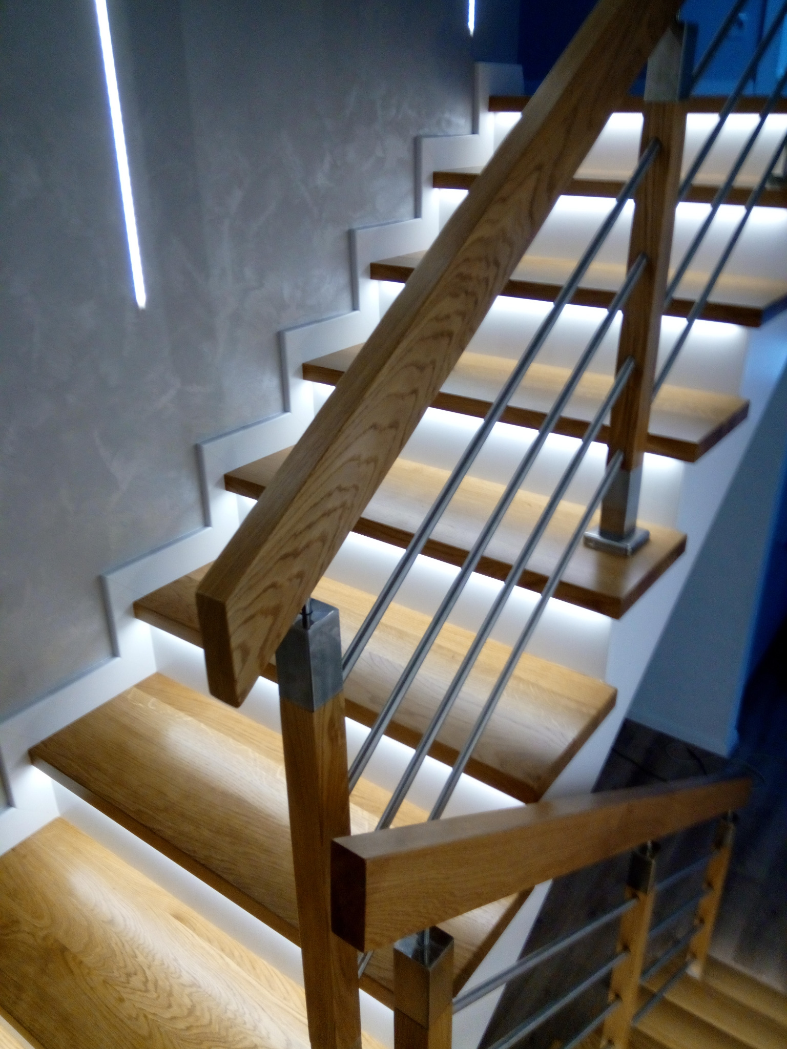 Nowoczesne schody z drewna z podświetleniem LED pod każdym stopniem i balustradą z poziomych, metalowych prętów.