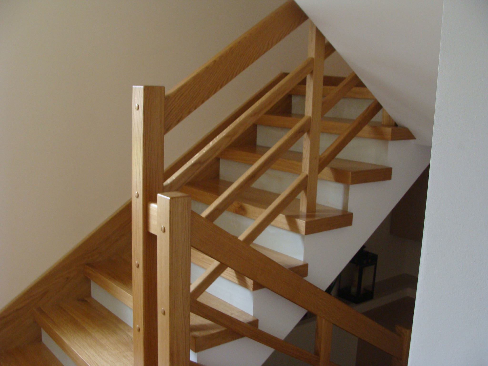 Jasne schody drewniane z prostą balustradą, widoczne mocowania słupków na gładkie nity, w tle fragment dekoracyjnej latarenki.