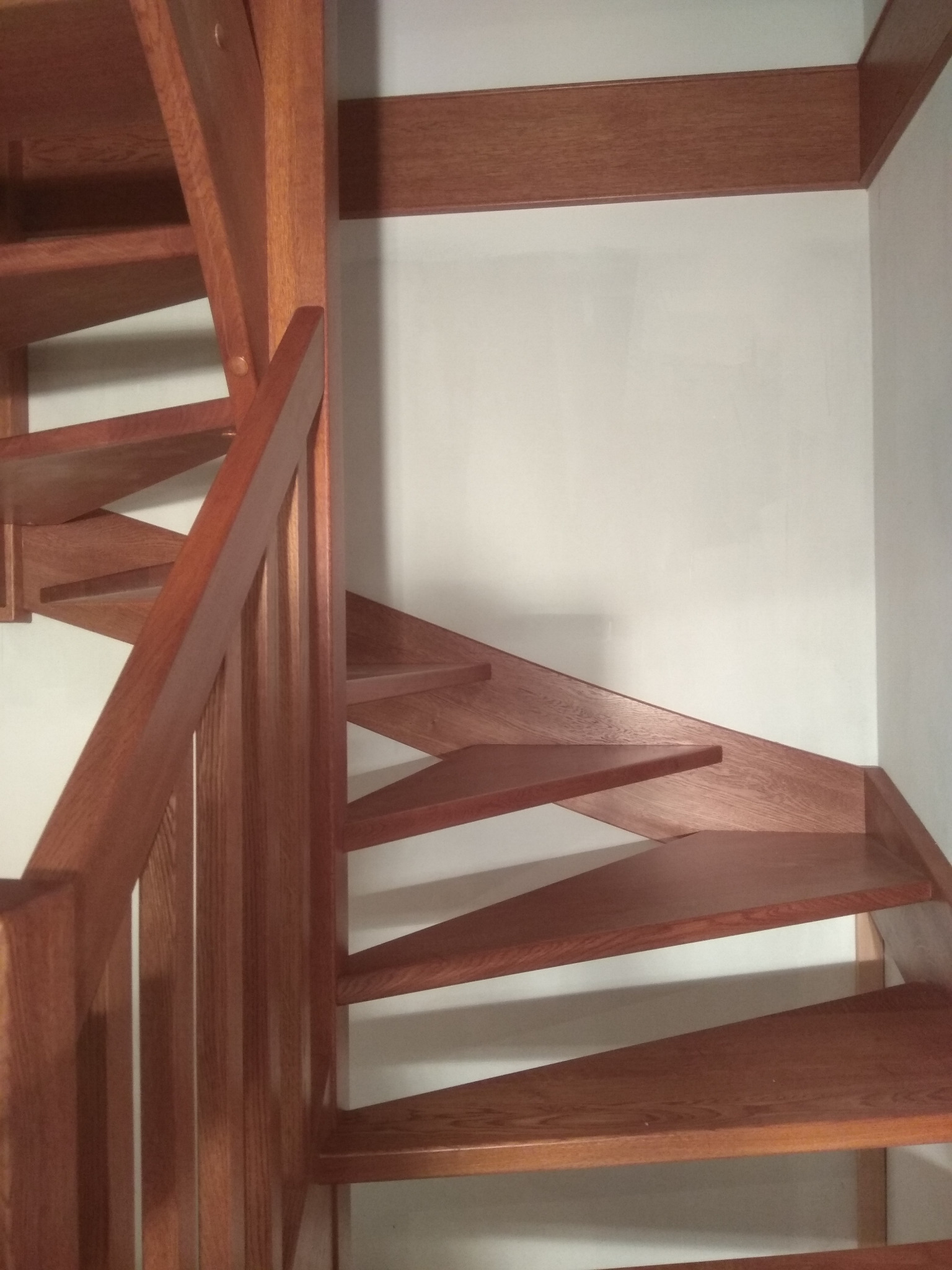 Kompaktowe, spiralne schody z drewna o ciepłym odcieniu, z widoczną strukturą drewna i minimalistyczną balustradą, w jasnym wnętrzu.