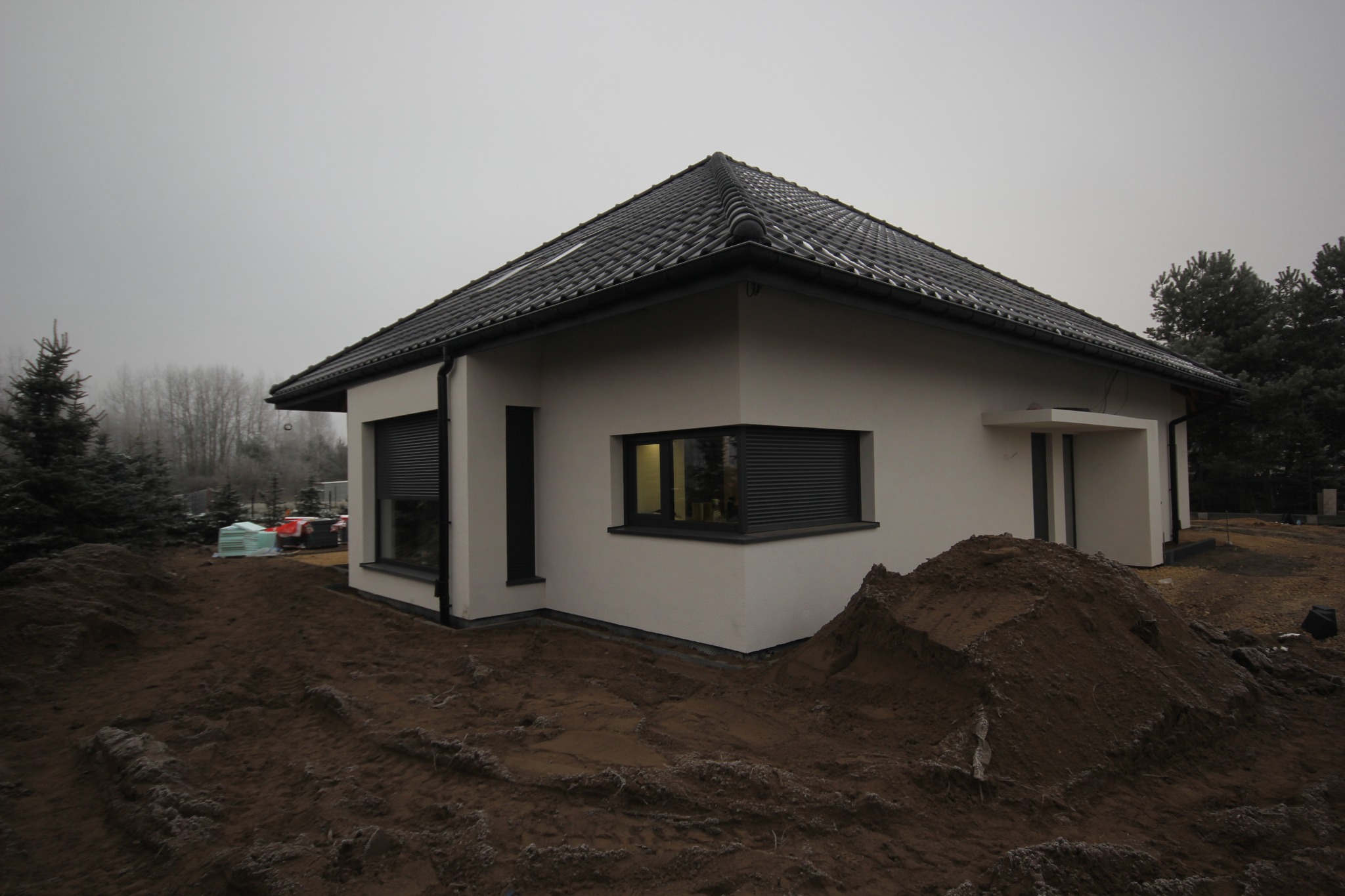 Nowoczesny dom jednorodzinny z czarną dachówką i oknami z zewnętrznymi roletami, otoczony ziemią przygotowaną pod ogród, widoczne materiały budowlane w tle.