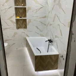Nowoczesna łazienka z wanną obudowaną drewnopodobną mozaiką w jodełkę, podświetleniem LED pod wanną, płytkami imitującymi marmur na ścianach i wnękami z drewnopodobnym wypełnieniem.
