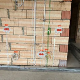 Surowa ściana z pustaków z widocznymi przewodami elektrycznymi, puszkami instalacyjnymi z czerwonymi zaślepkami, oraz korytem kablowym pod sufitem w trakcie montażu instalacji elektrycznej.